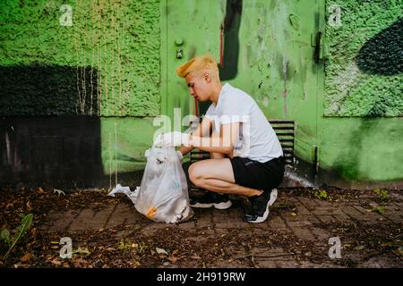 Vista laterale a tutta lunghezza del giovane maschio volontario accovacciato mentre si raccolgono rifiuti di plastica da parete verde Foto Stock