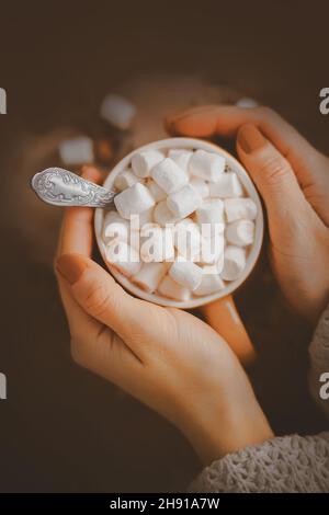 Una donna con un manicure marrone in un maglione di lana bianca sta tenendo una tazza beige con cacao caldo e marshmallow dolce. Una bevanda calda piacevole e deliziosa Foto Stock