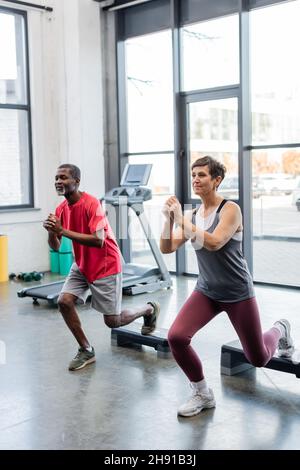 Donna anziana che si esercita con passi aerobici vicino afroamericano amico in palestra Foto Stock