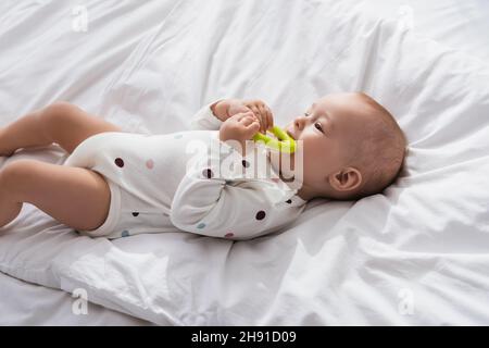 bambino bambino mordere il giocattolo mentre si trova su biancheria da letto bianca in romper Foto Stock