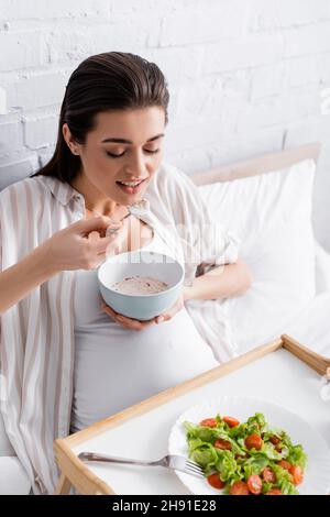 donna incinta felice mangiare farina d'avena vicino vassoio con insalata Foto Stock