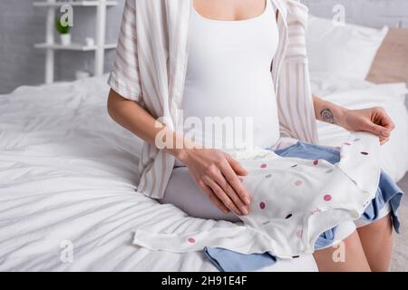 vista croppata della donna incinta tatuata che tiene i rompers del bambino nella camera da letto Foto Stock