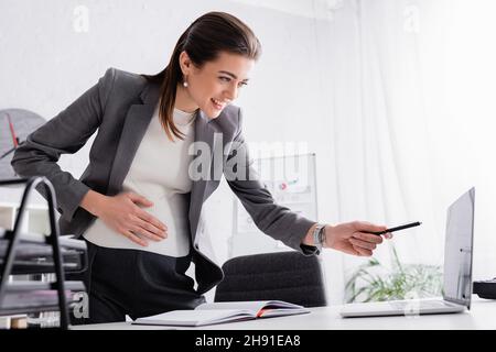 donna d'affari felice incinta che punta con la penna al laptop sulla scrivania Foto Stock