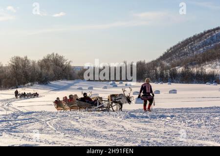 I turisti in una corsa in slitta con renne attraverso il paesaggio innevato in una soleggiata giornata invernale, guidata da un uomo Sami che indossa gákti (abito tradizionale) Foto Stock