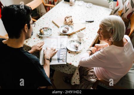 Vista ad angolo alto dell'infermiera maschile e della donna anziana che legge il giornale al tavolo da pranzo Foto Stock