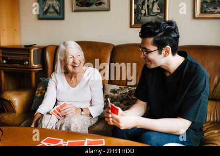 Happy maschio operatore sanitario giocare carte con la donna anziana in soggiorno Foto Stock