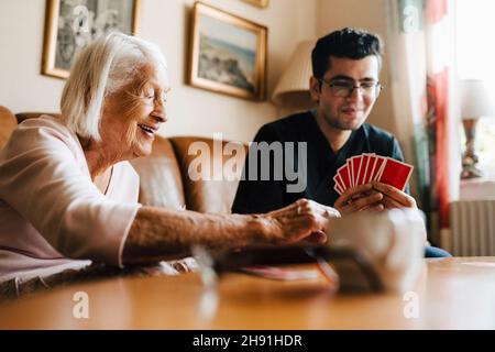 Donna anziana felice che gioca le carte con l'operatore sanitario maschile in soggiorno Foto Stock