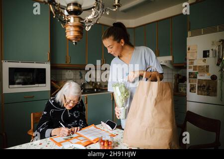 Giovane infermiere donna che guarda mentre donna anziana risolvere puzzle cruciverba nel libro a casa Foto Stock