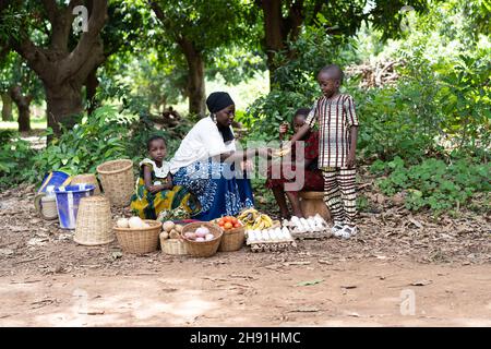 Carino ragazzo nero piccolo che acquista una banana da un gruppo di venditori di mercato di strada dell'Africa occidentale seduto sotto alcuni grandi alberi di mango sul lato della strada Foto Stock