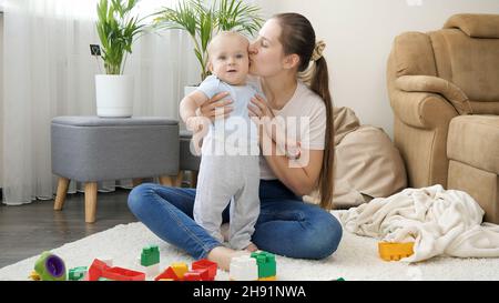 Felice sorridente madre seduta sul tappeto a casa e baciare il suo figlio del bambino Foto Stock