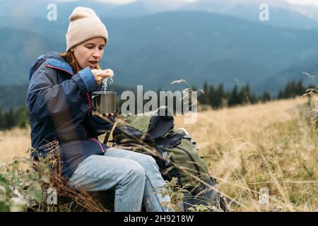 Ritratto di ragazza turistica caucasica mangiare fast food in montagna, spaghetti istantanei per il viaggiatore, delizioso pranzo in natura mentre rilassante. Foto Stock