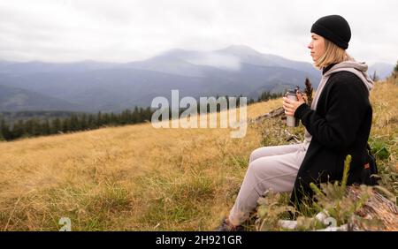 Una donna che viaggia in cima a un'alta montagna beve caffè da una tazza di metallo, ragazza guarda le montagne. Ragazza turistica libera, sfondo montagna Foto Stock