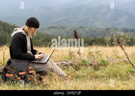 Donna caucasica che lavora sul suo computer in cima alla montagna. Concetto di lavoro remoto. Il viaggiatore lavora a distanza mentre si gode la montagna Foto Stock