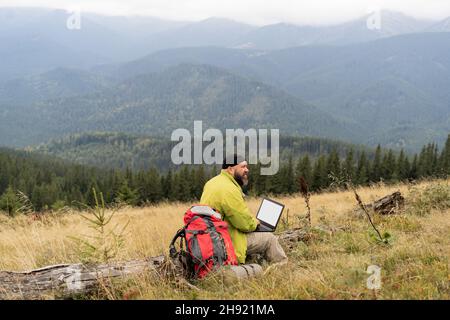 Un uomo caucasico barbuto con un computer portatile si siede su un albero caduto in montagna con una bella vista della valle. Foto Stock