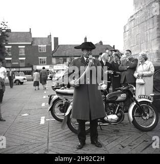 1965, storica, Ripon piazza della città, un hornblower guardato dai visitatori della città, casa di un antico Cathedal, Ripon, Yorkshire, Inghilterra, Regno Unito. Risalente al 886, un Wakeman ha tenuto sotto controllo la città di Ripon, assicurando che ci fosse qualcuno in guardia di notte. Ogni sera alle 9:00 per centinaia di anni, il soffiatore ha suonato il suo corno quattro volte intorno alla croce del mercato presso la piazza del mercato Ripon. Foto Stock