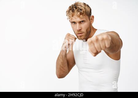 Bell'atleta maschile, pugilato ombra, pratica pugni di combattimento, estendi il pugno alla fotocamera, guardando focalizzato, sfondo bianco Foto Stock
