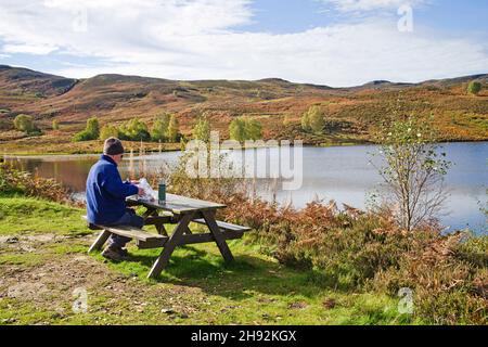Walker pranzare al tavolo da picnic da un piccolo lago collinare moorland nella soleggiata giornata autunnale, Tayside Sporting estate, Highland Perthshire, Scozia UK Foto Stock