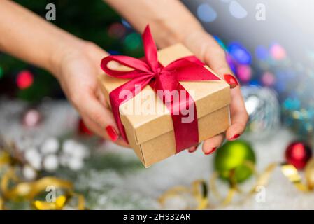 Confezione regalo con un arco nelle mani di una ragazza con una serpentina e giocattoli di Natale sullo sfondo di un albero di Natale e bokeh per il nuovo anno Foto Stock