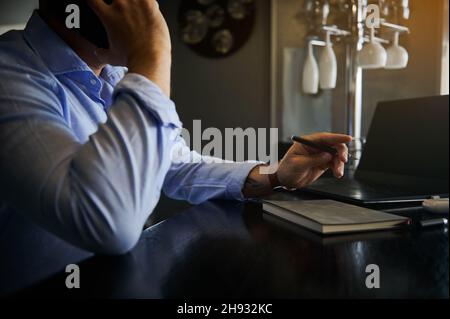 Concentrarsi sulla mano tenendo una penna di un uomo d'affari occupato e irriconoscibile, freelance parlare sul telefono cellulare mentre si lavora da remoto sul laptop a casa Foto Stock