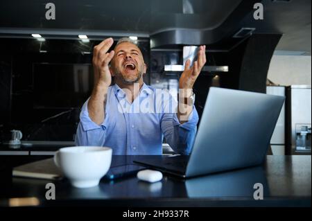 Bell'uomo d'affari di mezza età, imprenditore di successo solleva le braccia mentre gioisce e ride seduto di fronte al computer portatile alla scrivania con una tazza di fiuto Foto Stock