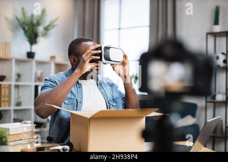 Uomo afroamericano che registra video sulla fotocamera mentre disimballa la scatola di cartone. Influencer maschile che offre recensioni online sui nuovi occhiali VR. Foto Stock