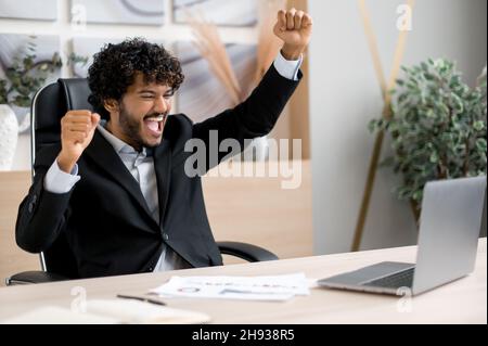 Riuscito gioioso eccitato uomo d'affari indiano, responsabile, commerciante di riserva, sieda ad un tavolo in ufficio, grida e gioisce al successo, alla vittoria o al buon profitto, guarda un laptop e i gesti con le sue mani Foto Stock