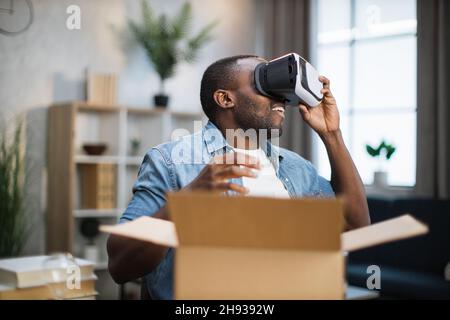 Happy african american man controllare il nuovo visore VR mentre si apre la confezione. Giovane blogger maschio che fa recensioni su nuovi prodotti per i suoi abbonati. Vista in prima persona. Foto Stock