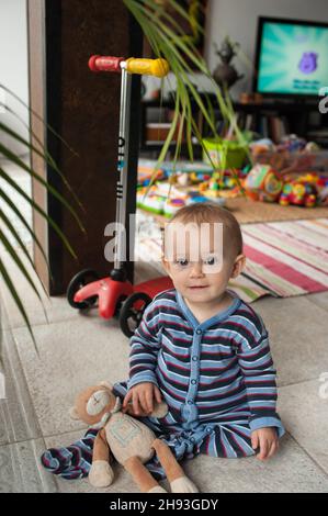 Una bambina (ca. 10 mesi) indossare un tuta stripy si siede nel soggiorno della sua casa circondato da giocattoli. Foto Stock