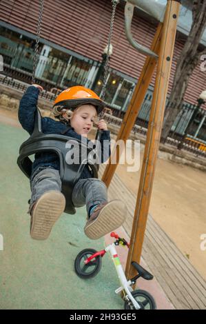 Un ragazzino (ca. 2 1/2 anni) gode di un giro su un altalena in un parco pubblico. Poggiando contro il telaio dell'altalena è la sua bicicletta apprendista senza pedali. Foto Stock