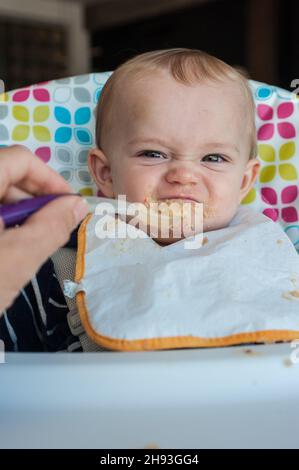 Una bambina (ca. 10 mesi) è stata poppata la sua cena di cibo di purea e rifiuta un cucchiaio con un grimace disgustato. Foto Stock