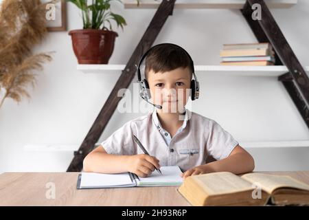 uno studente seduto con le cuffie su una scrivania ascolta l'insegnante e scrive i compiti in un notebook. Formazione online o tutor. Lezione di casa per un Foto Stock