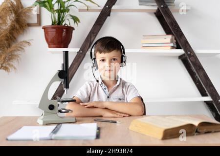 ragazzo studente in cuffie sta studiando a casa ha una lezione di biologia e un microscopio. Formazione online e insegnante di casa. Uno studente ascolta un webinar Foto Stock