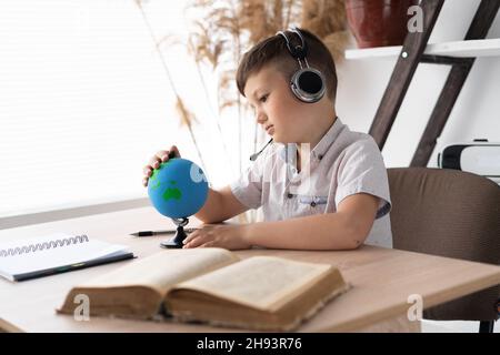 Un ragazzo triste che siede in cuffia ad una scrivania ascolta una lezione di geografia e guarda il mondo, studiando la contea e l'oceano. Wireless Foto Stock