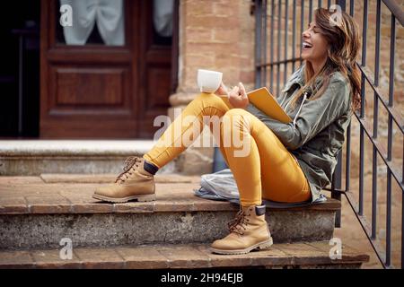 Una giovane ragazza è seduta sul portico di fronte all'edificio e godersi un libro e un caffè in un'atmosfera rilassata. Città, fuori, riposo Foto Stock