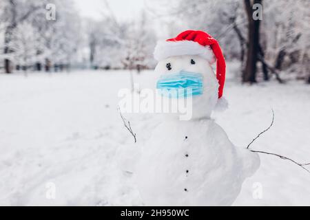 Snowman indossa maschera medica e cappello Santa nel parco invernale innevato. Misure protettive di sicurezza durante la pandemia di coronavirus covido Foto Stock