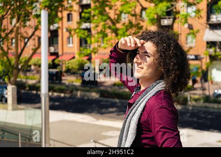 Un ritratto di un giovane uomo latino sorridente riccio che copriva gli occhi dal sole e che guardava al suo fianco con vecchi edifici sullo sfondo. Copia Foto Stock