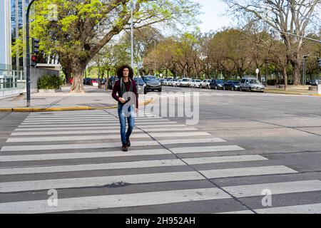 Un giovane uomo latino dai capelli ricci che attraversa la strada lungo il sentiero pedonale o zebra. Foto Stock