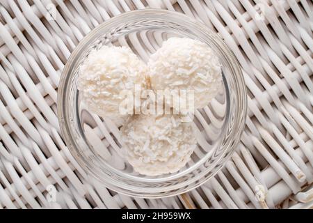 Tre caramelle bianche rotonde con scaglie di cocco in un piatto di vetro su un tappeto di vite, primo piano, vista dall'alto. Foto Stock