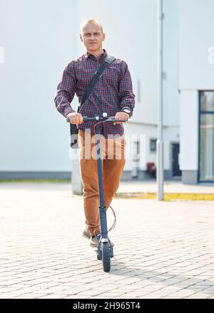 Giovane uomo in pantaloni casual e maglietta che guida il suo scooter elettrico, vista frontale, edifici sullo sfondo Foto Stock