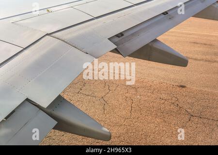 Vista dalla finestra dell'aeroplano commerciale, asfalto marrone sulla pista visibile sotto l'ala Foto Stock