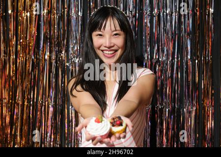 La giovane donna asiatica mostra i cupcake alla macchina fotografica sopra uno sfondo di festa. Foto Stock