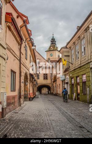 Un ciclista che scende lungo la strada di pietra di ciottoli/vicolo con un arco che porta a Piazza del Consiglio/brasov piazza principale (Piața Sfatului) in Brasov Romania. Foto Stock