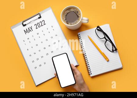 Mano femminile con telefono cellulare, appunti con calendario cartaceo per MAGGIO 2022, tazza di caffè, occhiali, penna e notebook su sfondo a colori Foto Stock