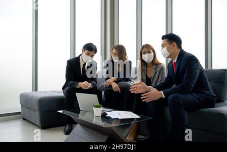 Gruppo di uomini d'affari in tuta discutere dei dati sul computer portatile. Tutti indossano la maschera facciale. Concetto di team aziendale di persone. Foto Stock