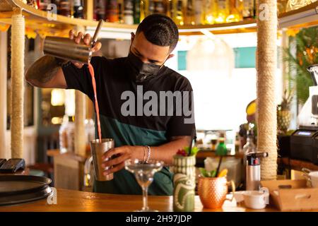 Giovane barista Latino che prepara un cocktail al bar. Indossa una maschera medica. Foto Stock