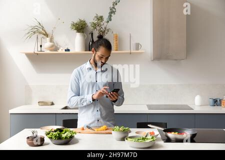 Felice uomo afroamericano usando il cellulare, cucinando in cucina. Foto Stock