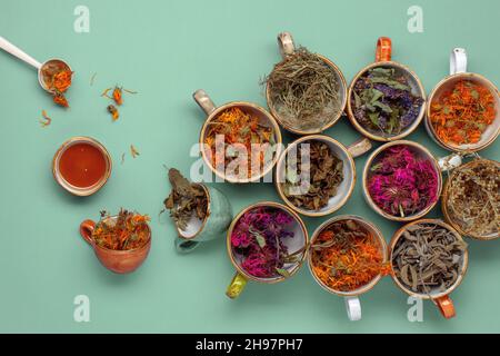 Assortimento di erbe di tè secche e rilassanti in tazzine colorate su sfondo verde menta con miele. Calendula, menta, issopo di anice, mondarda didima, legno di wormwood, Foto Stock