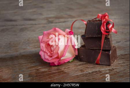 Pezzi di cioccolato e fiori su sfondo di legno. Tre pezzi di cioccolato con nastro rosso e rosa su superficie di legno scuro Foto Stock