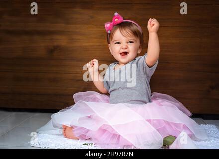 Il bambino urla con gioia. Ragazza bambino felice nella gonna rosa sullo sfondo di legno sollevato le mani e ride felicemente Foto Stock