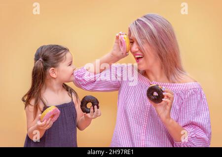 Divertente giovane donna e bambino su sfondo giallo parete. Madre e sua figlia ragazza si divertono con ciambelle colorate. Ciambelle rosa e al cioccolato. Foto Stock